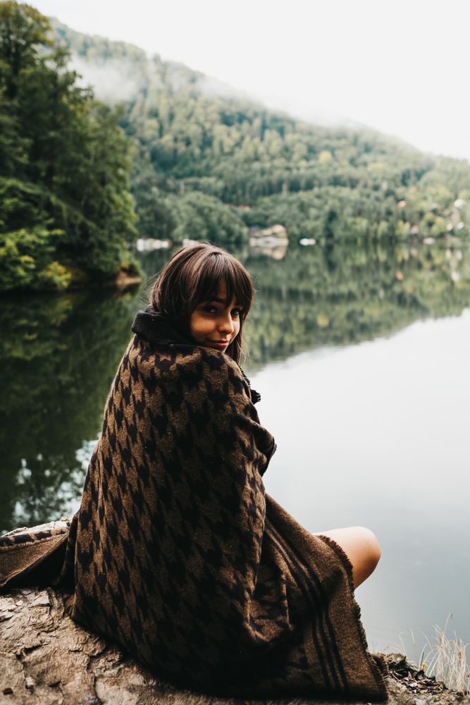 Woman sitting with blanket near lake and mountains