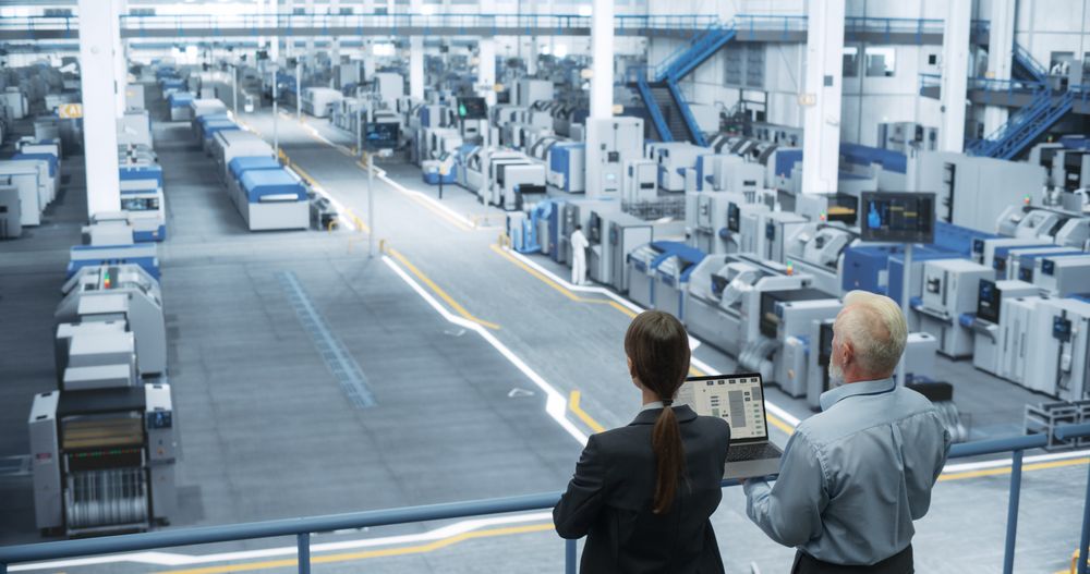 Male Engineer and Female Project Manager Standing on a Platform, Talking, Using Laptop Computer and Monitoring Production at a Modern Automated Electronics Manufacture with AI Robotic Arms