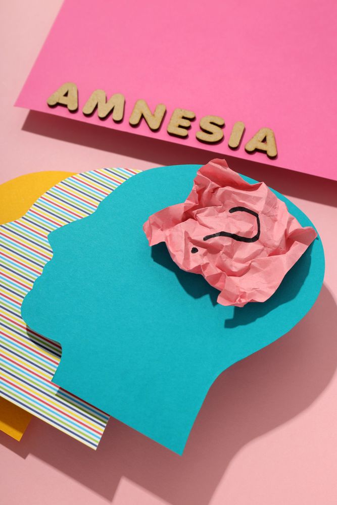 Vertical closeup of paper cut outs of colorful head on pink background with the word “Amnesia”