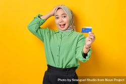 Confused Muslim woman in headscarf and green blouse holding up credit card beR260