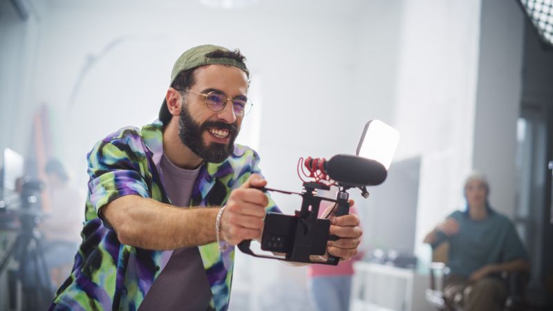 Young Male Videographer With Beard, Wearing Colorful Shirt And Cap, Joyfully Operates Camera Equipment In A Well-lit Studio, Surrounded By Creative Team Members Engaged In Film Production.