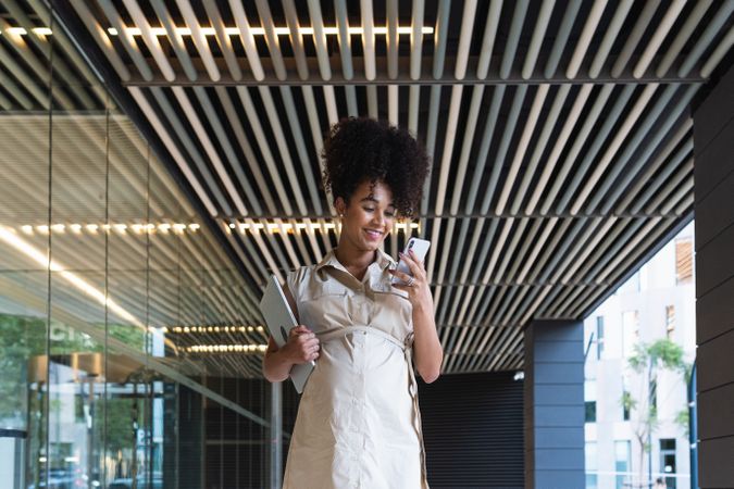 Smiling Black pregnant woman using smartphone and carrying laptop