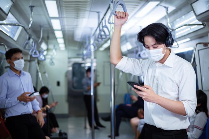 Male in facemask and headphones looking at smartphone in metro car