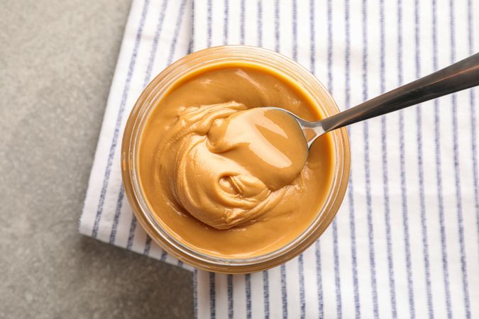 Glass jar with peanut butter and spoon, and kitchen towel on grey background, space for text and top view
