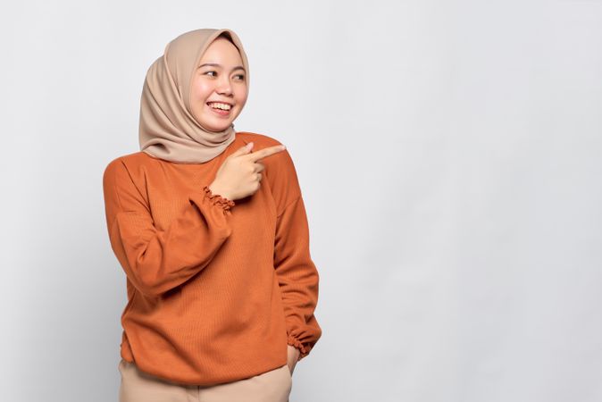 Smiling Muslim woman in headscarf and orange sweater pointing to copy space on her side