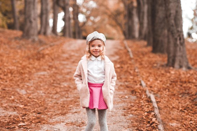 Young girl wearing pink jacket and gray hat standing on brown tree leaves