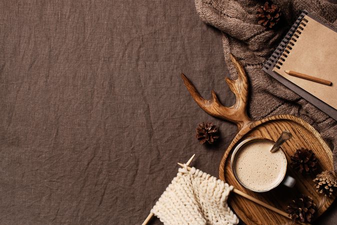 Overhead view of cozy cup of hot chocolate, knitting supplies, notebooks on textured brown fabric