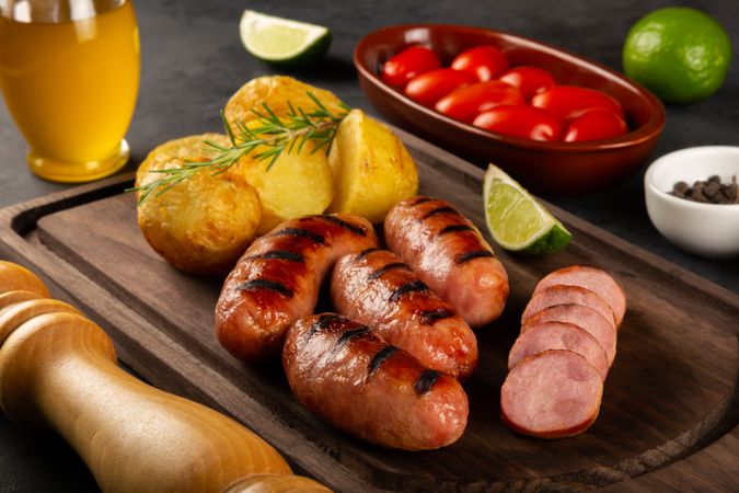 Wooden board with grilled sausages, potatoes, lime slices. tomatoes and sprig of rosemary on table