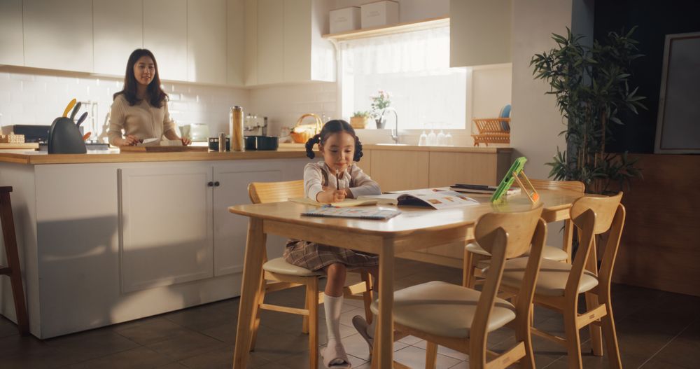 Portrait of a Korean Female Child Sitting at a Kitchen Table and Drawing While her Mother is Preparing Breakfast.