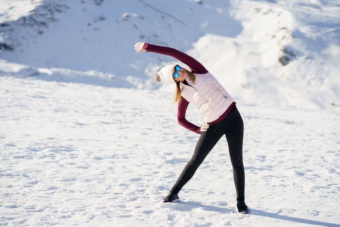 Full body of female in hat, polarized sunglasses, stretching sideways on on snowy mountain