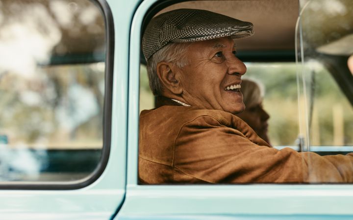 Side view of smiling mature man wearing hat driving a vintage car with woman sitting at the back