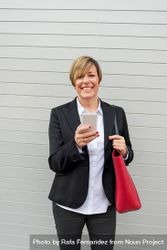 Smiling businesswoman holding out smartphone in front of wall 0yRQR5