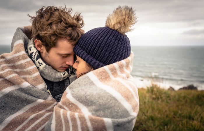 Closeup of young couple embracing under blanket in a cold day with ocean and dark cloudy sky in the background