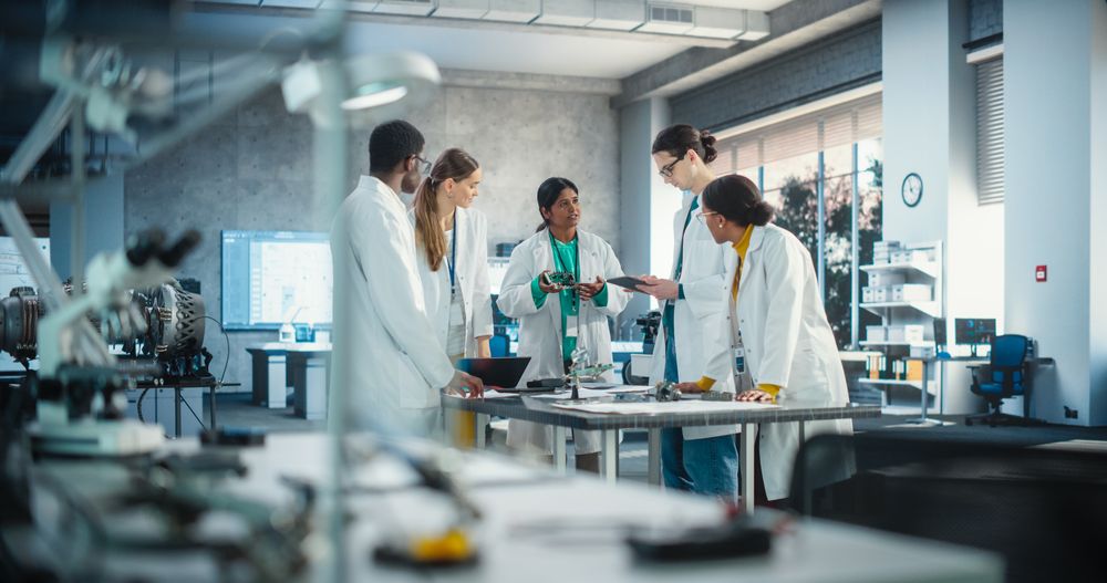 Group of International Multiethnic Lab Specialists Gathering Around a Table to Discuss Blueprints and Components for a High Tech Robotic Project.