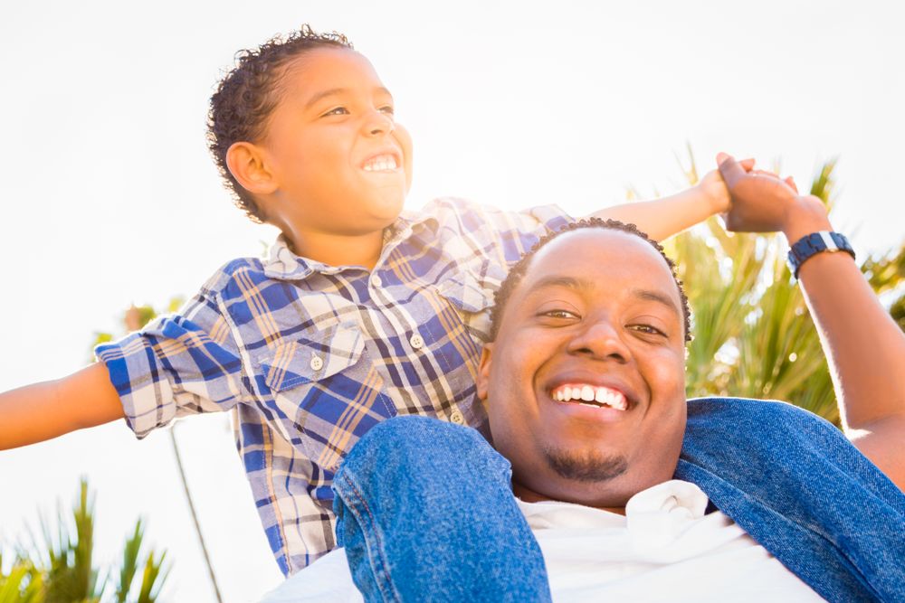 Happy Son Piggybacking On Father's Back Outdoors Together On Sunny Day