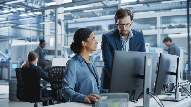 Portrait of a Young Male Team Leader and Hispanic Female Engineer Collaborating on a Project, Using Computer and Talking in a Factory with Robotic Arms Producing Modern Devices for Tech Industries.