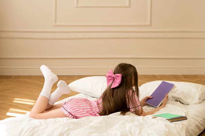 Girl in pajamas with pink bow reading book on bed