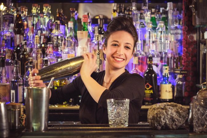 Cheerful female bartender mixing cocktail in shaker at counter