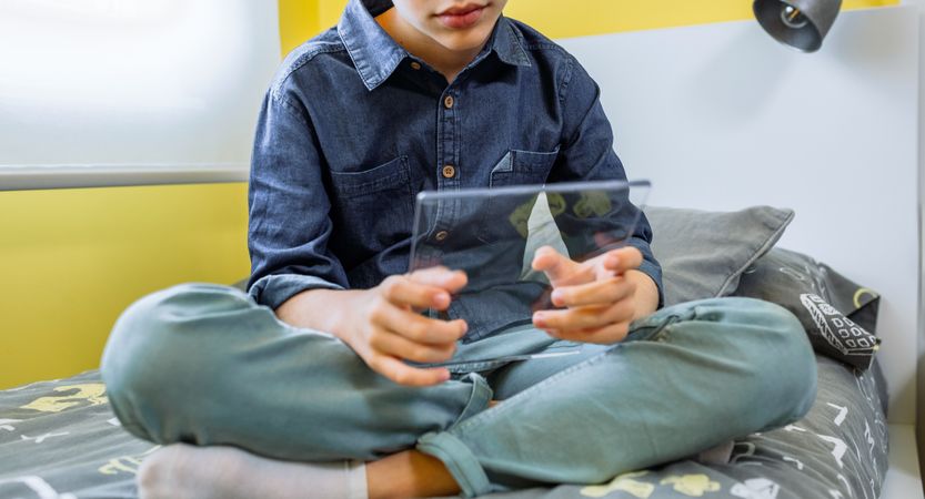 Unrecognizable boy using futuristic transparent tablet sitting over his bed