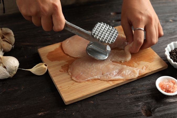 Hands of cook tenderizing chicken breast on board with mallet