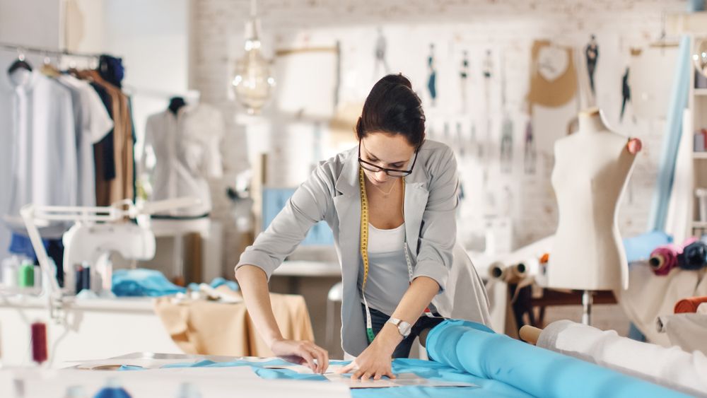 Brunette fashion, designer,  Lays Out Templates on the fabrics and Looks at Her Smartphone.