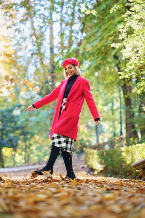 Full body of happy female in red clothes and beret walking on pathway with withered leaves in autumn park with trees