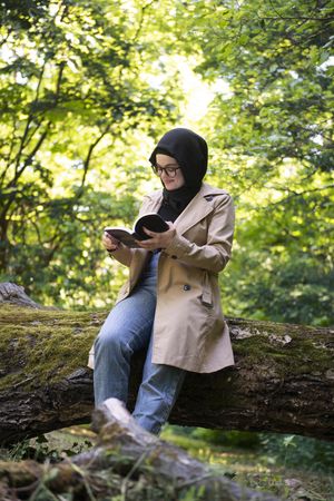 Happy Middle Eastern woman sitting on mossy tree with book
