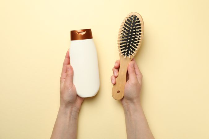 Female hands hold shampoo bottle and hair brush on beige background