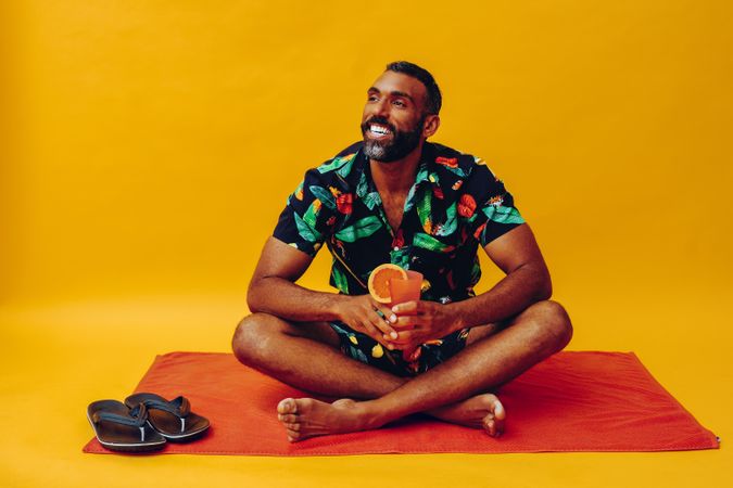 Black male sitting on towel and looking away while holding up tropical drink