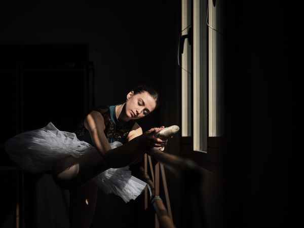 Young ballerina stretching leg on barre in dark room