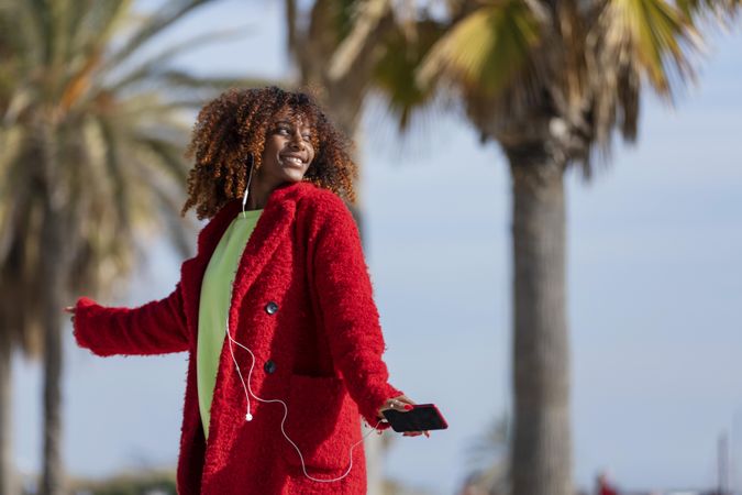 Happy female turning around while listening to music on phone on street with palm trees