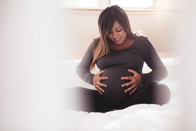 Pregnant woman touching her belly with hands while sitting on bed at home