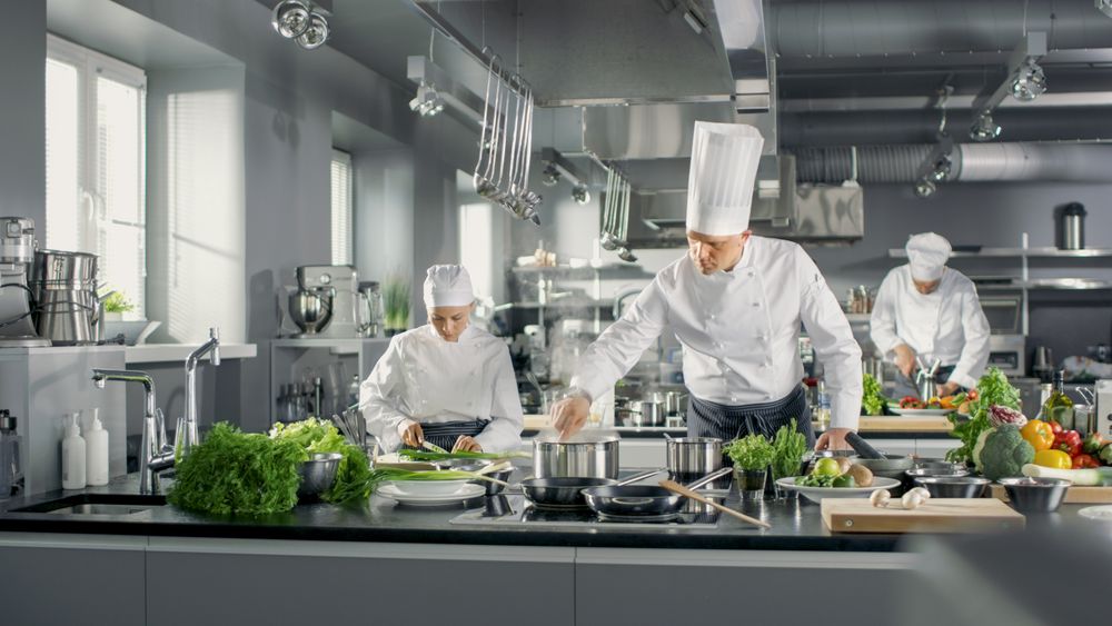 Famous Chef Works in a Big Restaurant Kitchen with His Help. Kitchen is Full of Food, Vegetables and Boiling Dishes.