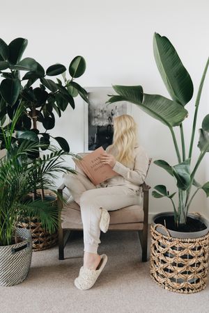 Blonde woman reading a book sitting on a chair between indoor plants