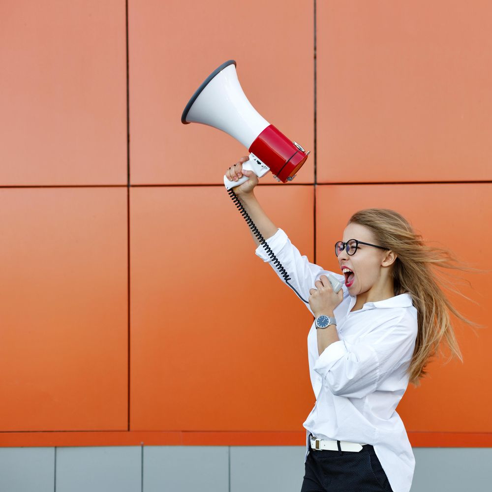Woman in glasses in front of orange wall speaking into megaphone, square