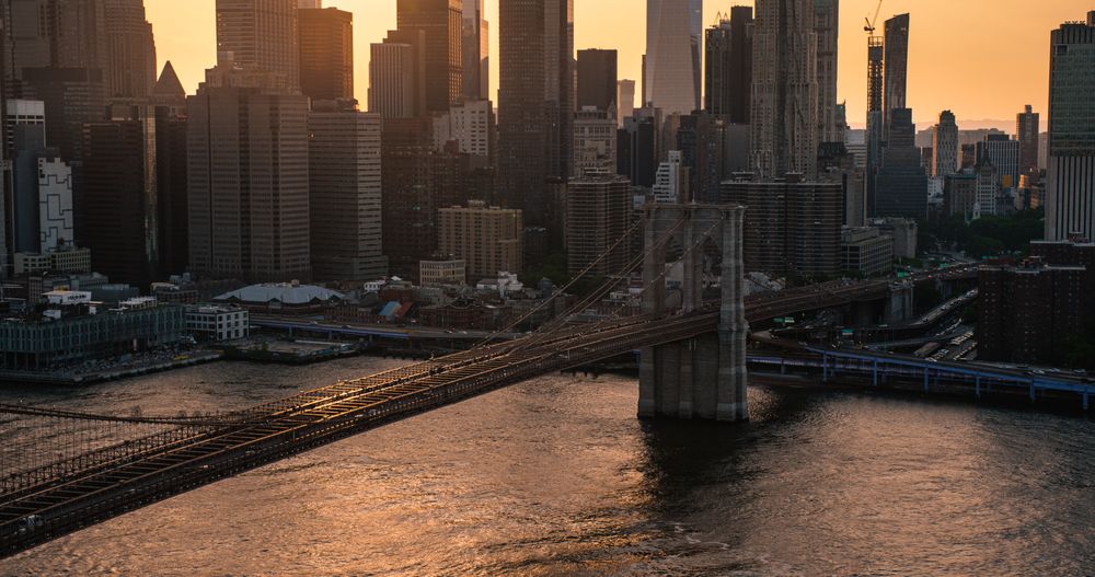 Iconic New York City Landscape Over East River with Skyscrapers, Brooklyn Bridge, Cars and Ferry Boats. Cinematic Evening Urban Skyline with Sunset and Lightly Clouded Sky