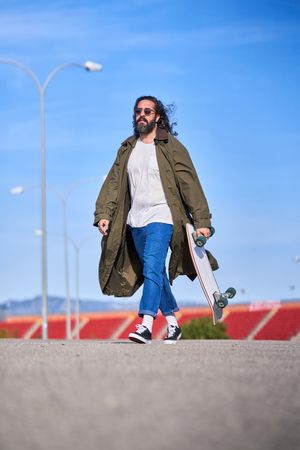 Trendy hipster man in raincoat walking with skateboard