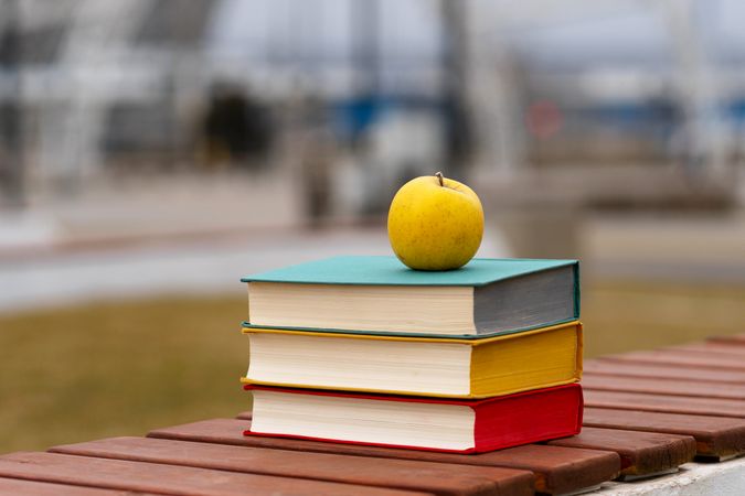 Yellow apple placed atop stack of hardcover books outdoors.
