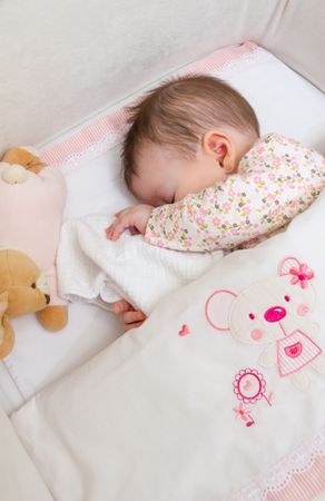 Baby girl covering her face, asleep in a cot with pacifier and toy