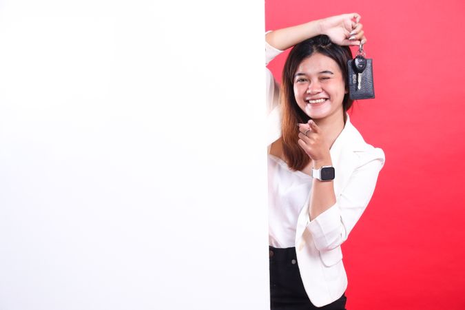 A smiling woman with brown hair is holding a car key and pointing at the camera, she is wearing a jacket, isolated on a red background