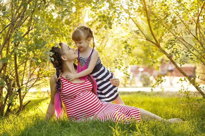 Pregnant female in red striped dress smiling at her daughter under the trees while sitting on grass