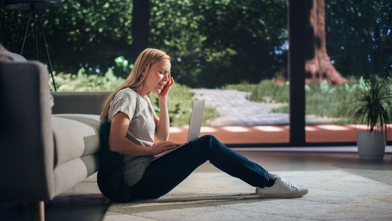 Portrait of a Young Adult Female Experiencing Sudden Migraine while Sitting on a Floor and Using Laptop Computer in Sunny Living Room.