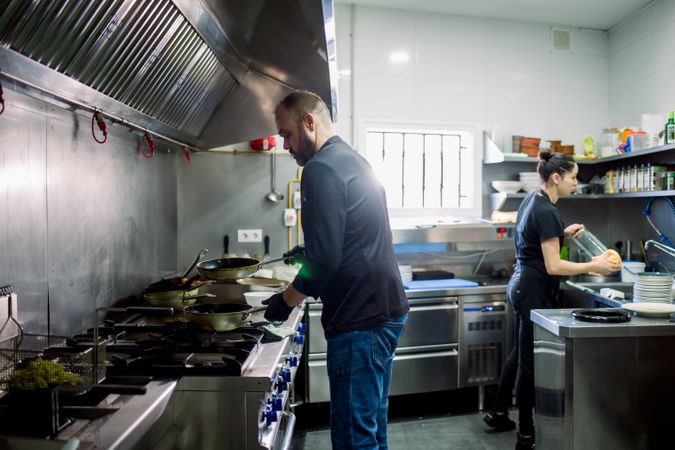 Side view of bearded male chef and woman cooking food over pan and washing dishes in modern kitchen of restaurant