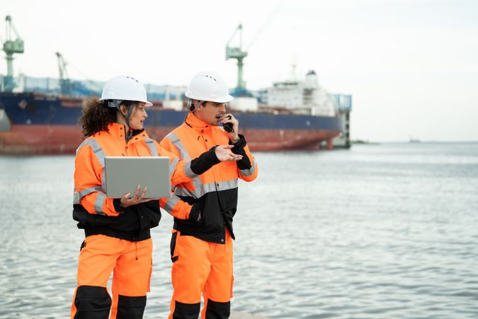 Workers in safety helmets examine laptop screen at sea, Freight ship in background, Dockyard engineers collaborate by harbor, man uses walkie-talkie, Port facility teamwork, Logistics industrial