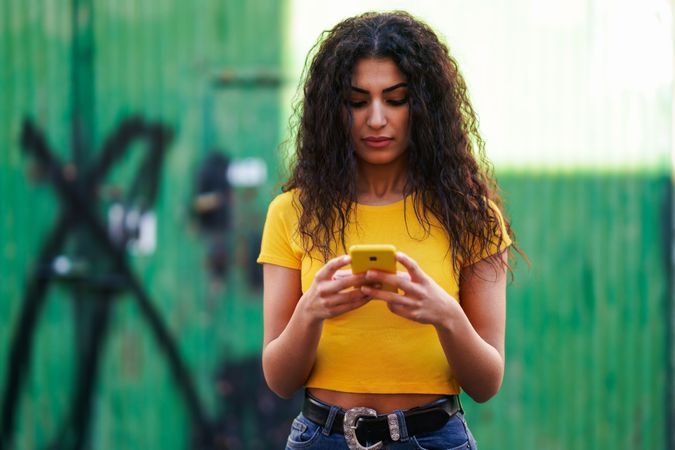 Woman with curly hair in yellow t-shirt and jeans texting on phone, copy space