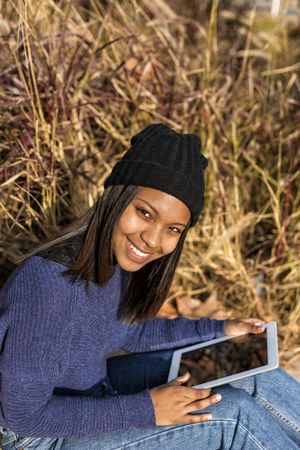 Smiling female in hat and sweater sitting in grass with tablet