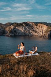 Woman with glass of drink on picnic mat looking at sea and mountains