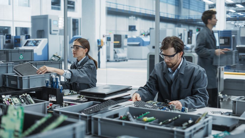 Male And Female Workers Taking Apart Old Laptops To Recycle Electronic Components For Printed Circuit Board Production At Electronics Factory.
