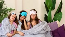 Three girls with eye masks checking temperature, sitting on couch.