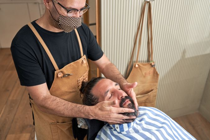 Crop barber in mask massaging chin of male customer